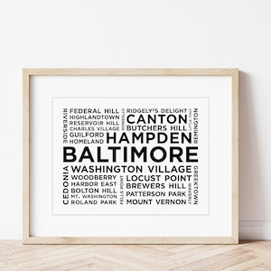 May include: A framed print featuring the word "Baltimore" in bold black text, surrounded by the names of its neighborhoods in a minimalist design. The print is in a light wooden frame, perfect for home decor.