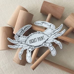 May include: A black and white illustration of a crab with the text "LOCUST POINT" on its shell. The illustration is on a sticker and is placed on top of several wooden mallets.
