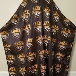 Puede incluir: Una capa de barbero negra con un patrón repetido de logotipos de cabeza de jaguar dorados con la palabra "JAGS" impresa dentro de cada logotipo.