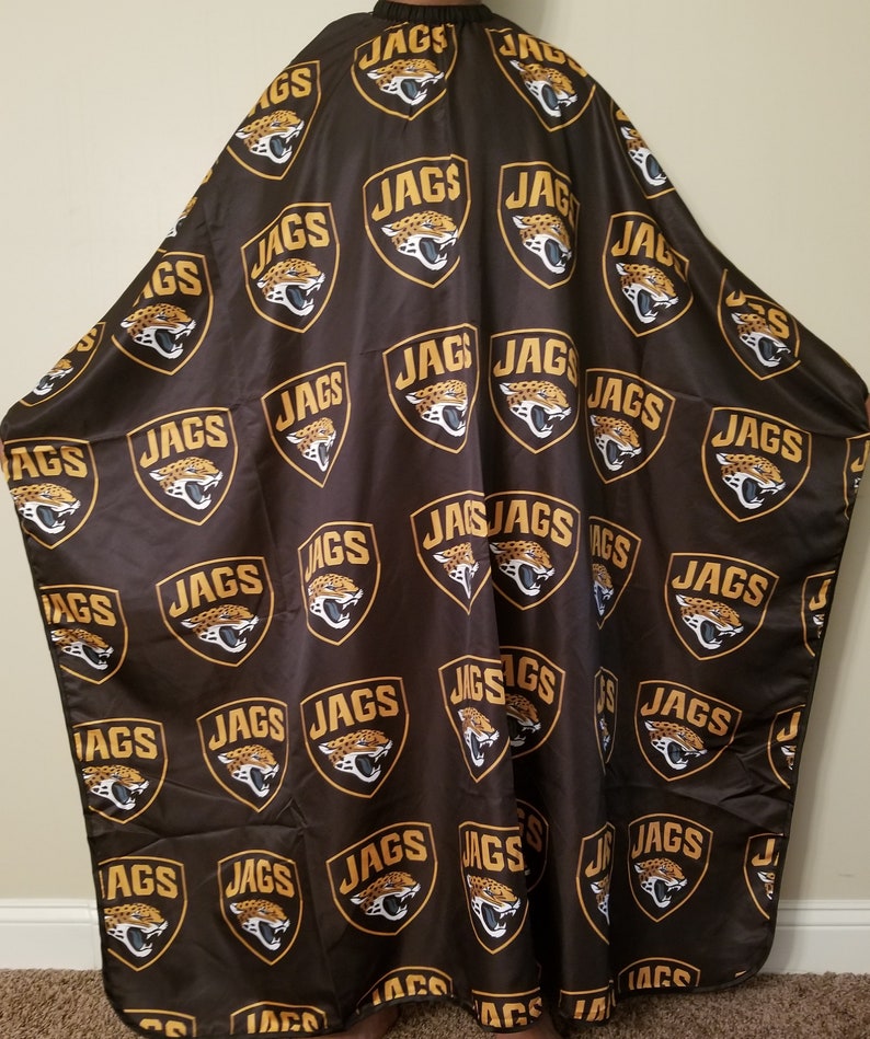 K&ouml;nnte beinhalten: Eine schwarze Friseurumhang mit einem sich wiederholenden Muster aus goldenen Jaguar-Kopflogos mit dem Wort "JAGS" in jedem Logo.