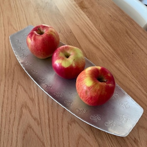 Könnte beinhalten: Drei runde rote und gelbe Äpfel, die in einer Reihe auf einem länglichen, rechteckigen silberfarbenen Metalltablett angeordnet sind. Das Tablett weist ein sich wiederholendes geprägtes Blumenmuster an den Rändern auf.