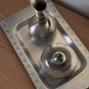 May include: A silver-toned metal serving tray featuring a repeating floral border pattern, accompanied by a matching metal pitcher with a textured neck and a lidded container with a textured circular lid design.