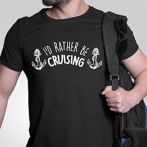 Könnte beinhalten: Ein schwarzes T-Shirt mit weißem Text, der "I'd rather be cruising" lautet, mit zwei Anker-Symbolen auf jeder Seite des Textes.