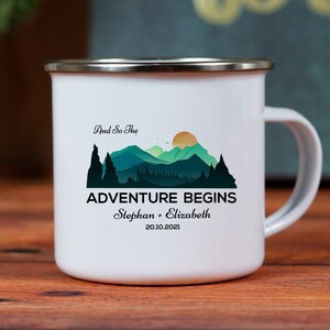Op de afbeelding: Witte emaille campingmok met een berglandschap en de tekst "And So The Adventure Begins" met de namen "Stephan + Elizabeth" en de datum "20.10.2021".