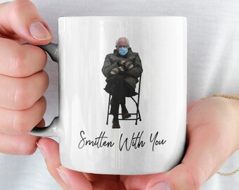 May include: White ceramic mug with a photo of a person wearing a face mask and sitting on a chair. The text "Smitten With You" is printed on the mug.