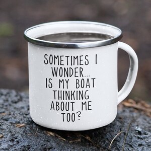 May include: White enamel camping mug with a silver rim and handle. The mug has black text that reads "SOMETIMES I WONDER... IS MY BOAT THINKING ABOUT ME TOO?" The mug is speckled and sits on a dark, wet surface.