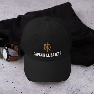 May include: Black baseball cap with a gold embroidered ship's wheel and the text "CAPTAIN ELIZABETH" in white.
