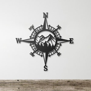 May include: Black metal compass wall decor featuring a mountain silhouette design. The compass includes the cardinal directions and a decorative border.