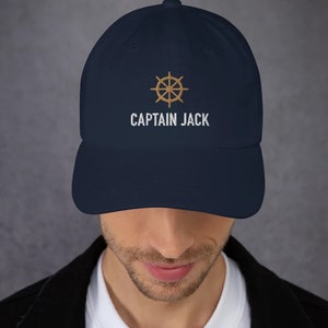 May include: A navy blue baseball cap with a gold embroidered ship's wheel and the text "CAPTAIN JACK" in white.