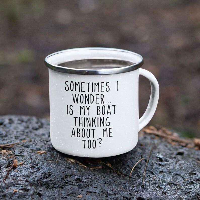 May include: White enamel camping mug with a silver rim and handle. The mug has black text that reads "SOMETIMES I WONDER... IS MY BOAT THINKING ABOUT ME TOO?" The mug is speckled and sits on a dark, wet surface.