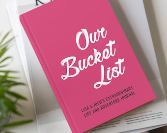 May include: A hot pink journal titled "Our Bucket List" with white script lettering. The journal also includes the text "Lisa & Josh's Extraordinary Life and Adventure Journal". The journal is resting on top of a magazine.