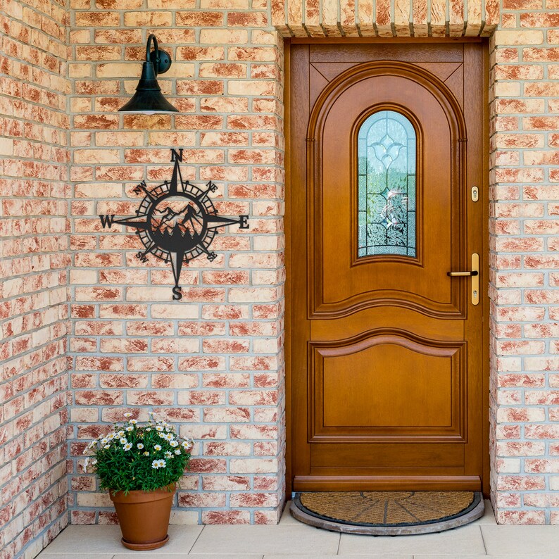 May include: Black metal compass with mountain silhouette design mounted on a brick wall next to a wooden door with a glass window.