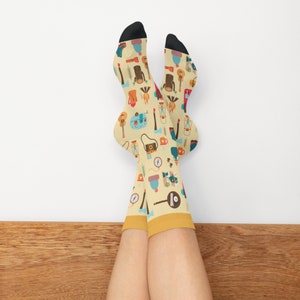 May include: A pair of yellow socks with a colorful pattern of camping and outdoor adventure themed icons. The socks are on a person's legs, with the feet crossed.