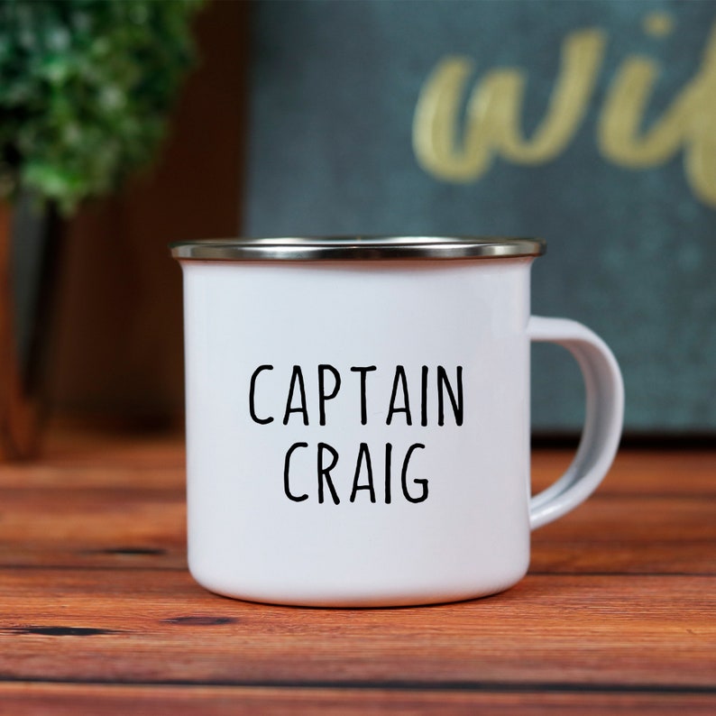 May include: White enamel mug with a silver rim and a handle. The mug features the words "CAPTAIN CRAIG" in black, bold letters. The mug is on a wooden surface.