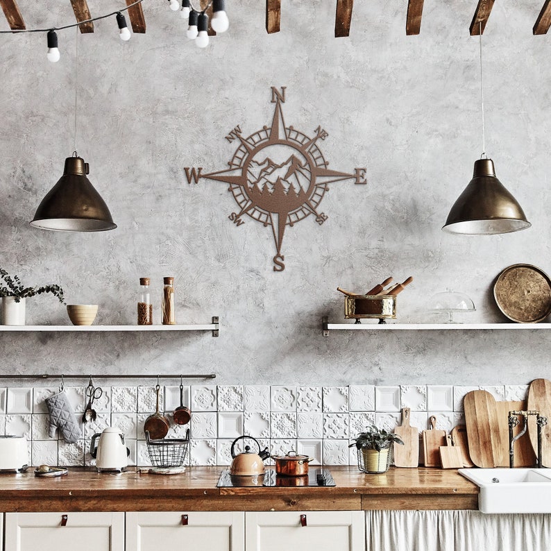 May include: A brown metal compass with a mountain scene inside the compass rose. The compass is mounted on a grey wall in a kitchen setting.