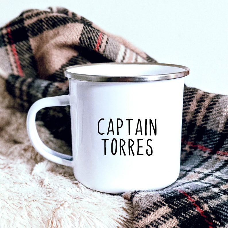 May include: White enamel mug with a silver rim and a black handle. The mug features the text "CAPTAIN TORRES" in black, centered on the front. The mug is set against a backdrop of a plaid blanket and a fluffy, cream-colored surface.