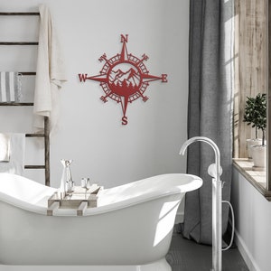 May include: Red metal compass wall decor featuring a mountain and tree design. The compass points north, south, east, and west.