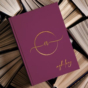 May include: A burgundy journal with a gold circle and the letter "A" inside. The name "ashley" is written in gold script below the circle.