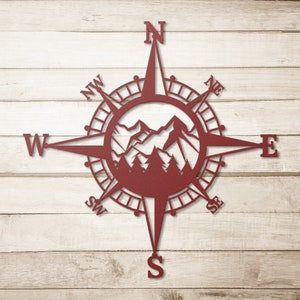 May include: A red metal compass with a mountain range design in the centre. The compass points north, south, east, and west. The compass is on a wood background.