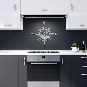 May include: A silver metal compass with a mountain scene inside the compass. The compass is mounted on a black wall in a kitchen setting.