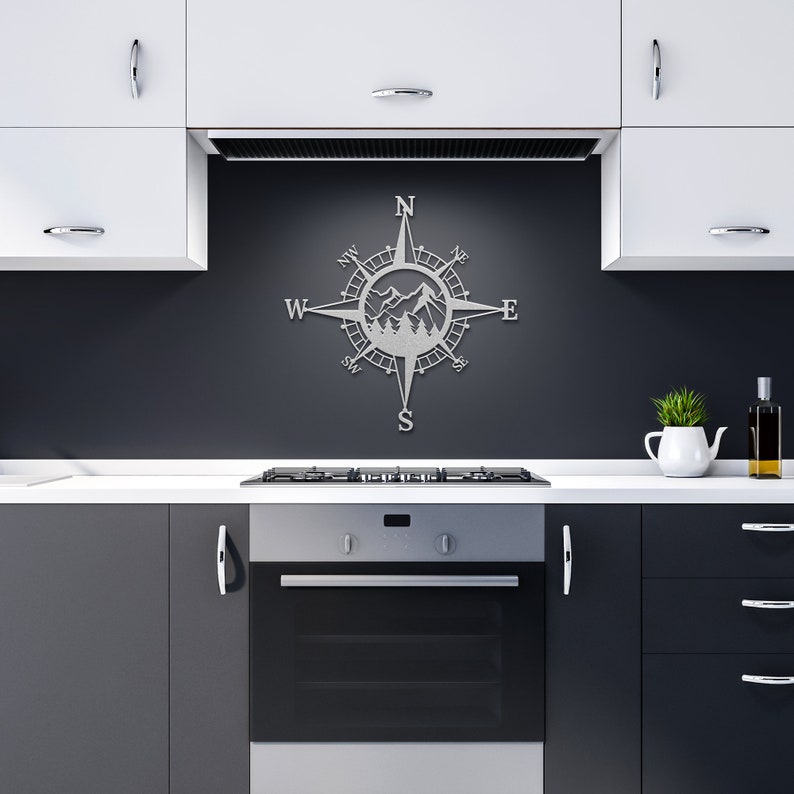 May include: A silver metal compass with a mountain scene inside the compass. The compass is mounted on a black wall in a kitchen setting.