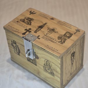 Puede incluir: Una caja de madera clara con un cierre metálico, adornada con imágenes religiosas grabadas y texto. La caja muestra ilustraciones de figuras, cruces y texto bíblico, incluyendo "PSALM 30:5" y "MATTHEW 6:6".