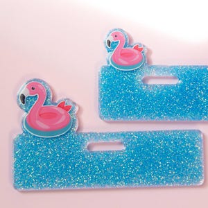 Puede incluir: Dos porta-credenciales azules brillantes con un diseño de flamenco rosa. El flamenco es rosa con un pico blanco y un acento rojo. Los porta-credenciales tienen forma rectangular con un recorte para un cordón.