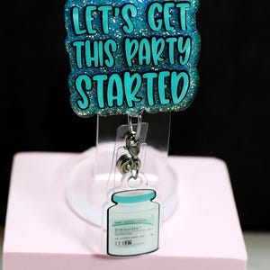 Könnte beinhalten: Ein Ausweis-Rollhalter mit dem Text "LET'S GET THIS PARTY STARTED" in teal und blauem Glitzer. Die Rolle hat einen transparenten Kunststoffclip und eine kleine, weiß-blaue Grafik einer Ampulle. Die Worte "Hot Mess Creative Shop" sind ebenfalls sichtbar.