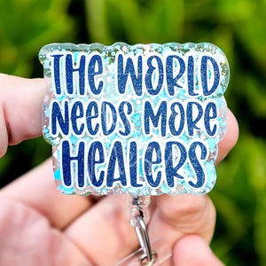 Può includere: Un portabadge con la scritta "The World Needs More Healers" in blu. Il badge ha un bordo trasparente e glitterato e una clip retrattile argentata. Lo sfondo è verde sfocato.