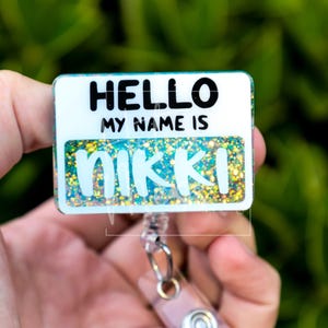 Pode incluir: Uma placa de identificação retangular com o texto "HELLO MY NAME IS NIKKI" em preto. O nome "NIKKI" está em uma fonte brilhante e colorida. A placa é presa a um carretel retrátil com um clipe prateado.