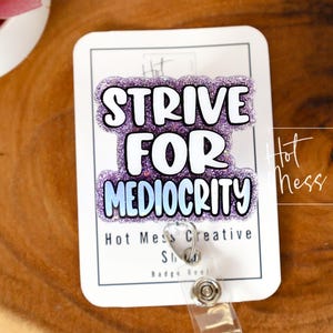 May include: A white badge reel with the text "STRIVE FOR MEDIOCRITY" in white and purple glitter. The badge reel has a clear plastic strap and a metal clip. The background is a wooden surface.