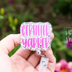 Puede incluir: Una insignia con las palabras "CERTIFIED YAPPER" en letras de color rosa brillante. La insignia tiene un borde plateado brillante y un clip de pl&aacute;stico transparente. El fondo est&aacute; borroso con follaje verde y rosa.