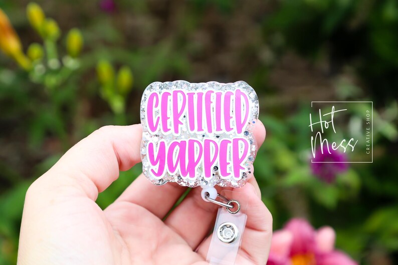Puede incluir: Una insignia con las palabras "CERTIFIED YAPPER" en letras de color rosa brillante. La insignia tiene un borde plateado brillante y un clip de pl&aacute;stico transparente. El fondo est&aacute; borroso con follaje verde y rosa.