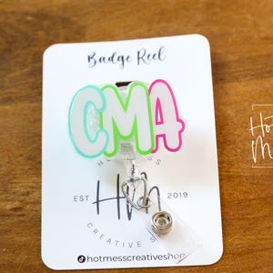 K&ouml;nnte beinhalten: Ein Ausweis-Rollhalter mit den Buchstaben "CMA" in einer Bubble-Schrift, mit einem Farbverlauf aus Hellblau, Gr&uuml;n und Pink. Die Rolle ist an einer wei&szlig;en Karte mit dem Text "Badge Reel" und "Hot Mess Creative Shop" befestigt.