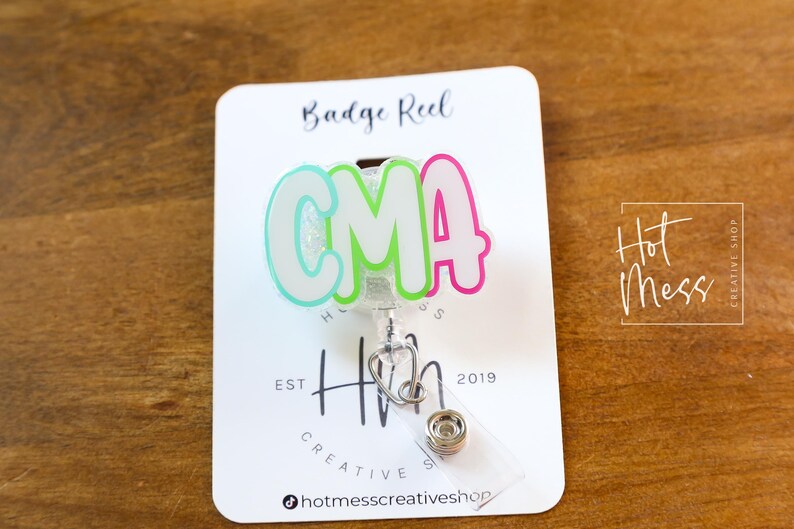K&ouml;nnte beinhalten: Ein Ausweis-Rollhalter mit den Buchstaben "CMA" in einer Bubble-Schrift, mit einem Farbverlauf aus Hellblau, Gr&uuml;n und Pink. Die Rolle ist an einer wei&szlig;en Karte mit dem Text "Badge Reel" und "Hot Mess Creative Shop" befestigt.