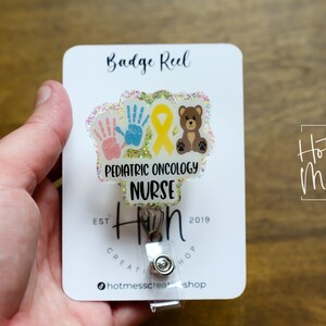 Peut inclure: Un porte-badge transparent avec un fond paillet&eacute; et un motif repr&eacute;sentant deux empreintes de mains, un ruban jaune et un ours en peluche brun. Le texte "Pediatric Oncology Nurse" est imprim&eacute; sur le porte-badge. Le porte-badge est attach&eacute; &agrave; une carte blanche avec le texte "Badge Reel" et "Hot Mess Creative Shop" imprim&eacute; dessus.