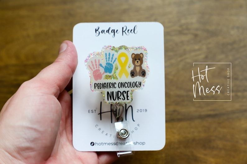 Peut inclure: Un porte-badge transparent avec un fond paillet&eacute; et un motif repr&eacute;sentant deux empreintes de mains, un ruban jaune et un ours en peluche brun. Le texte "Pediatric Oncology Nurse" est imprim&eacute; sur le porte-badge. Le porte-badge est attach&eacute; &agrave; une carte blanche avec le texte "Badge Reel" et "Hot Mess Creative Shop" imprim&eacute; dessus.