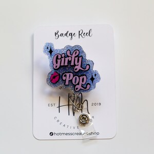 Op de afbeelding: Een badge reel met een glinsterend blauw en roze ontwerp met de tekst "Girly Pop" en een roze lip-afbeelding. De badge reel is bevestigd aan een witte kaart met de tekst "Badge Reel" en het logo van de winkel.