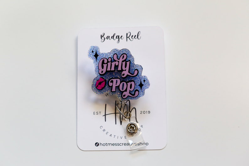 Op de afbeelding: Een badge reel met een glinsterend blauw en roze ontwerp met de tekst "Girly Pop" en een roze lip-afbeelding. De badge reel is bevestigd aan een witte kaart met de tekst "Badge Reel" en het logo van de winkel.