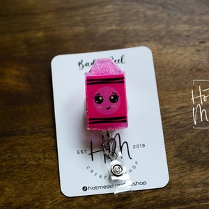 May include: Pink glitter badge reel with a cartoon crayon design. The crayon has a smiling face with big eyes. The text "Hot Mess Creative Shop" is on the card.