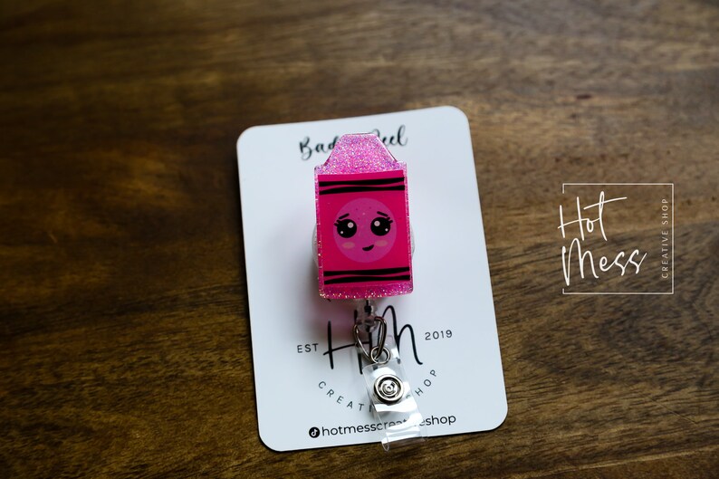 May include: Pink glitter badge reel with a cartoon crayon design. The crayon has a smiling face with big eyes. The text "Hot Mess Creative Shop" is on the card.