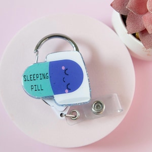 Op de afbeelding: Een badge reel met een slaappil ontwerp. De pil is wit, blauw en teal, met de woorden "SLEEPING PILL" gedrukt op het teal gedeelte. Het blauwe gedeelte heeft een slapend gezicht met gesloten ogen. De reel heeft een zilveren clip en een doorzichtige plastic band.
