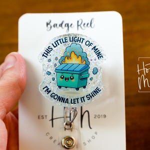May include: A white badge reel with a glitter-covered, clear plastic charm featuring a cartoon trash can with a smiling face and flames coming out of it. The charm has the text "This little light of mine, I'm gonna let it shine".