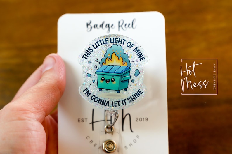 May include: A white badge reel with a glitter-covered, clear plastic charm featuring a cartoon trash can with a smiling face and flames coming out of it. The charm has the text "This little light of mine, I'm gonna let it shine".