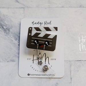 May include: A white card with black text that reads "Badge Reel" and a black and white cartoon clapperboard with a smiling face. The clapperboard is attached to a silver retractable badge reel. The card also has the text "Hot Mess Creative Shop" and "Est. 2019".