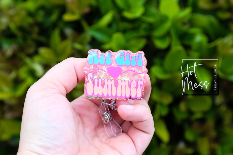 Pode incluir: Porta-crach&aacute; de resina rosa e azul com glitter e o texto "Hot Girl Summer" e um cora&ccedil;&atilde;o rosa.