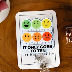 Puede incluir: Un portatarjetas blanco con una escala de caras emoji que representan emociones, de feliz a triste. El texto "IT ONLY GOES TO TEN" se muestra debajo. El portatarjetas está sobre una superficie de madera.
