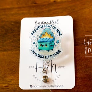 May include: A white glitter badge reel with a blue and green cartoon trash can with a face and flames coming out of it. The text on the badge reel says "This little light of mine I'm gonna let it shine".