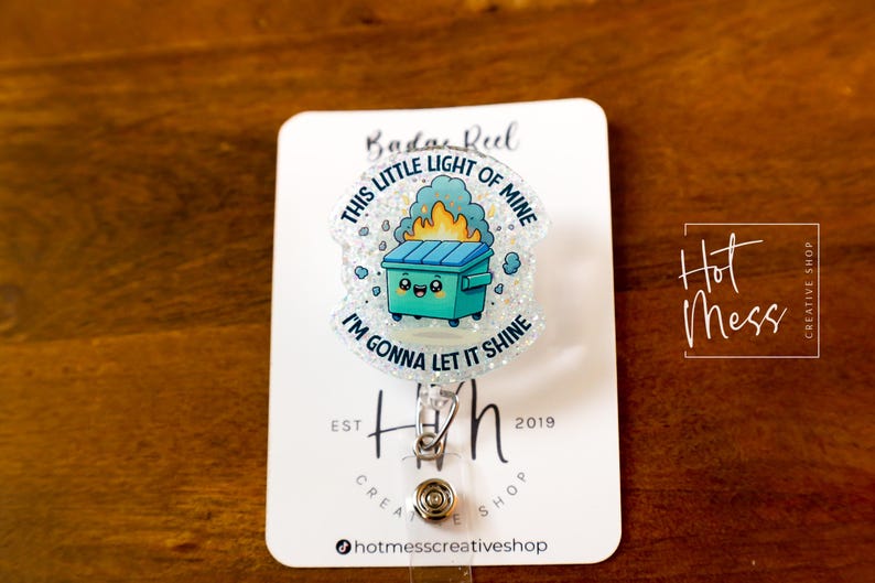 May include: A white glitter badge reel with a blue and green cartoon trash can with a face and flames coming out of it. The text on the badge reel says "This little light of mine I'm gonna let it shine".