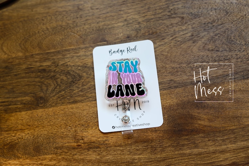 May include: A pink, blue and black glitter badge reel with the text "Stay in your lane" and the logo "Hot Mess Creative Shop" printed on a white card.